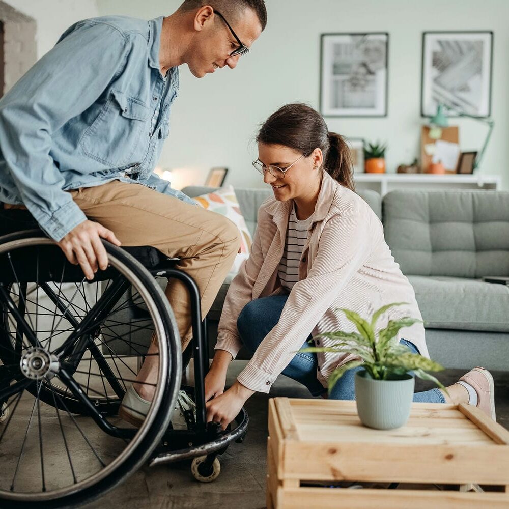 Spouse assisting partner who uses a wheelchair with daily activities at home, illustrating gratuitous family-provided care